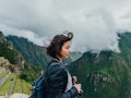A young woman stands in front of Machu Picchu in Peru with a camera around her neck.