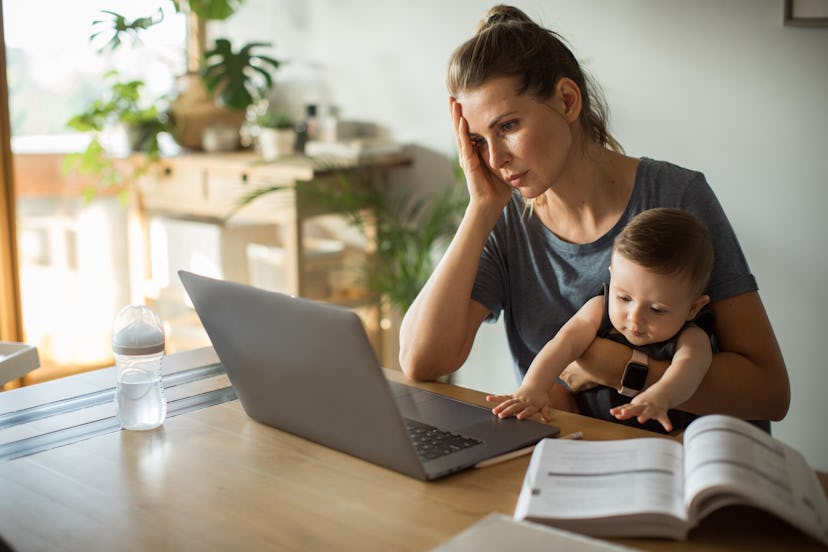 A person holds her baby at a table with a textbook propped open next to her laptop. The stress and a...