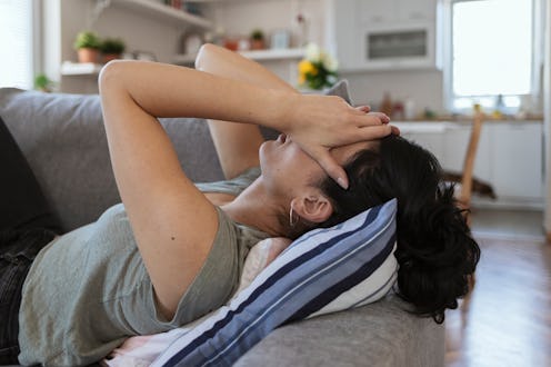 A person lays on her couch, propped up with pillows, with her head in her hands. If you're experienc...