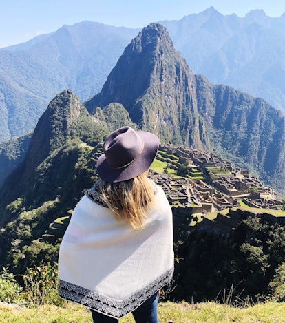 A young woman with blonde hair stands in front of Machu Picchu in Peru on a sunny day.