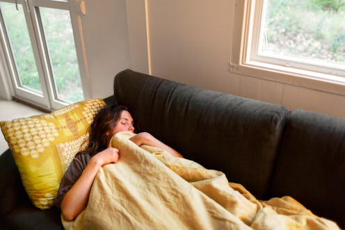 A woman takes a nap on a couch under a yellow blanket. If PMS or your period make you unusually tire...