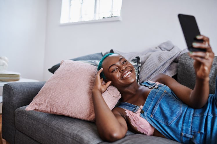 A young woman lays on a gray couch and records an Instagram Live video on her phone.