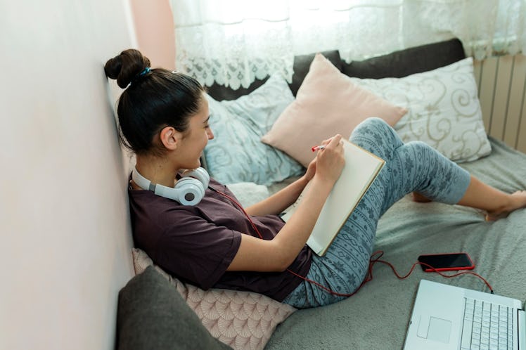 A young woman sits in her bed and draws a picture on a notepad while on video chat.
