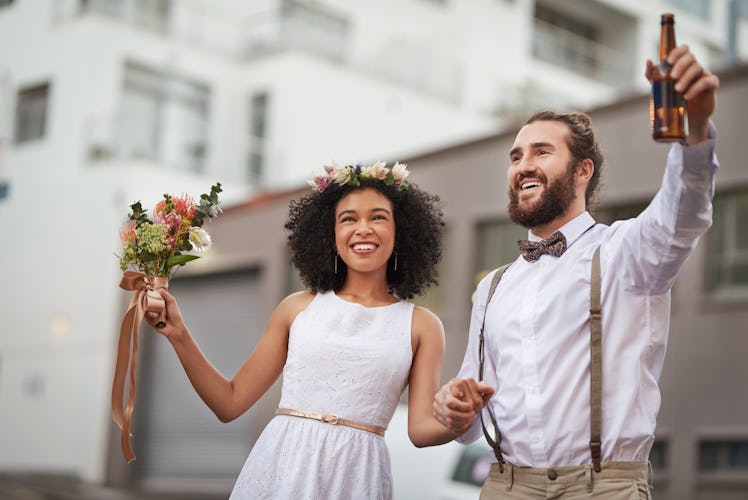 A young couple poses for a wedding photo in the city while holding a bouquet and bottle of beer.