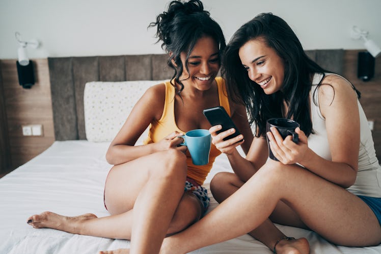 A young lesbian couple sits in bed with cups of coffee and texts on their phone.
