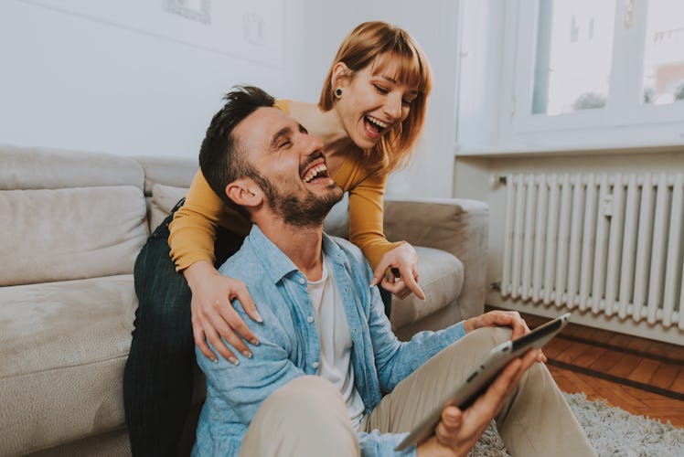 A young couple laughs while holding an iPad and hanging out in their cozy home.