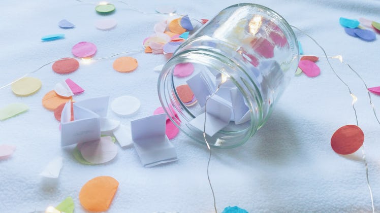 A mason jar is filled with folded paper and surrounded by string lights and confetti.