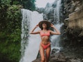 A young woman in a coral bathing suit poses near a rushing waterfall while on a trip.