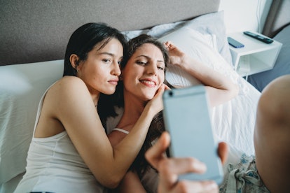 A young lesbian couple snuggles in bed and takes a selfie on a phone.