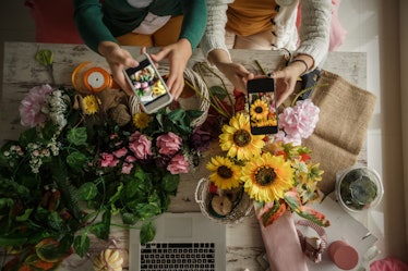 Two women sit at a table with bouquets of different flowers and a laptop.