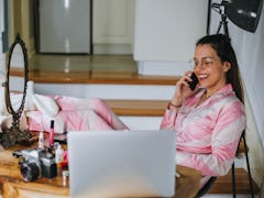 A young woman sits at a table with her laptop, a mirror, and makeup, and talks on the phone.