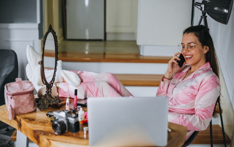 A young woman sits at a table with her laptop, a mirror, and makeup, and talks on the phone.