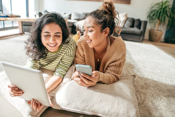 Two friends lay on the floor of their apartment and video chat on a tablet.