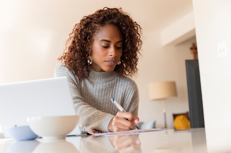 A young woman sits at her kitchen table with her laptop and jots things down on a piece of paper.