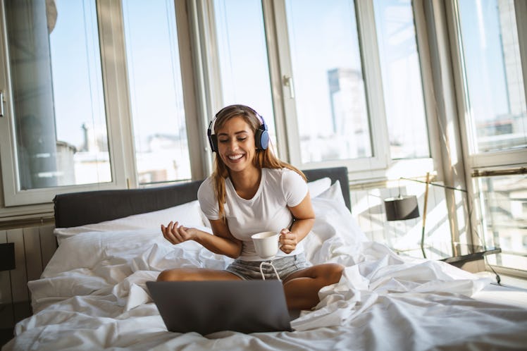 A young woman sits with her laptop and a cup of coffee in bed, while doing a virtual escape room and...