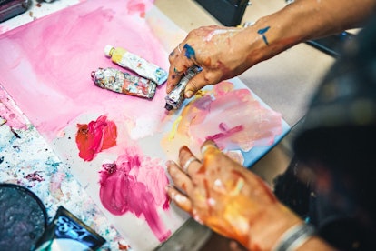 A young woman plays with watercolor paints on a paint-splattered table.