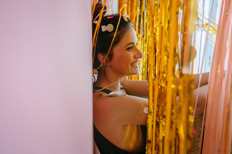 A young woman poses against a pink wall amongst gold streamers and confetti.
