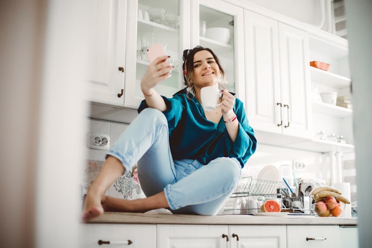A young woman sits on her kitchen counter with a cup of coffee and takes a selfie on her phone.