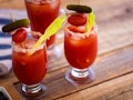 Two Bloody Marys placed on a picnic table have sticks of celery, and skewers of tomato, bacon, and s...