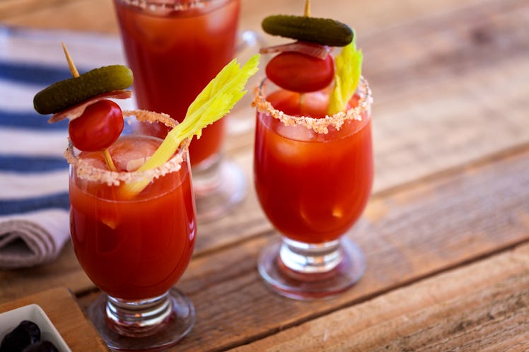 Two Bloody Marys placed on a picnic table have sticks of celery, and skewers of tomato, bacon, and s...