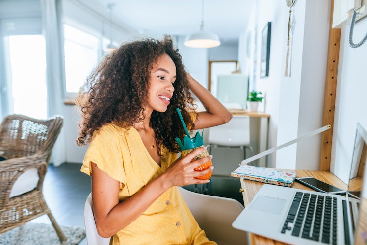 A young woman in a yellow dress sits at a desk with a tropical drink for virtual happy hour.