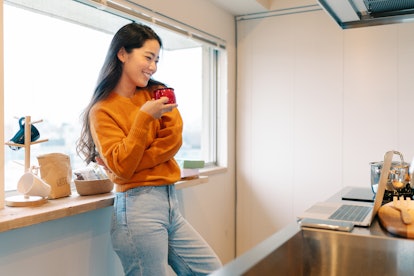 A young woman stands in her kitchen with a red mug while on video chat.