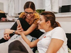 Two sisters sit on the couch and laugh at something on their phone. 