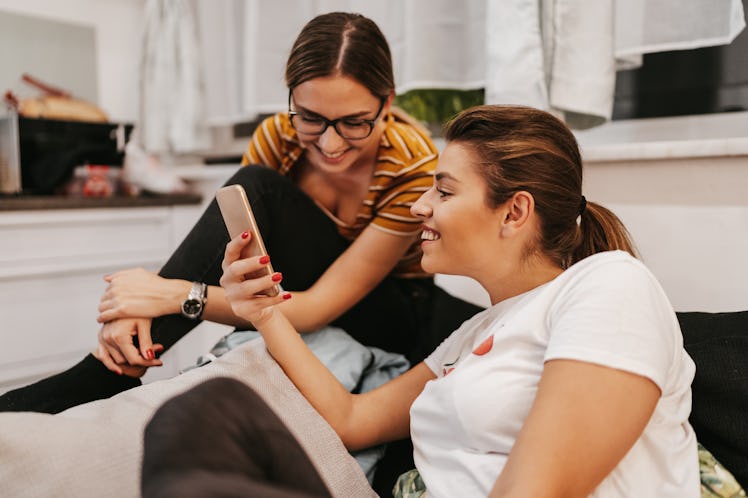 Two sisters sit on the couch and laugh at something on their phone.