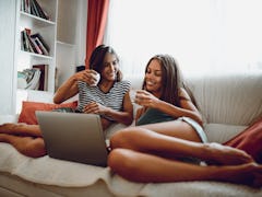 Two best friends sit on the couch and video chat with their other friends on a laptop. 