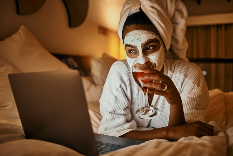 A young woman sits on her bed in a robe, does a face mask, and drinks wine while video chatting on h...