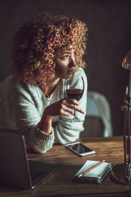 A young woman sits at her kitchen table with a glass of wine and her laptop.