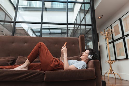 A woman with headphones watches Instagram live while lying on a brown velvet couch. Social distancin...