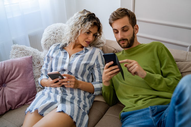 A young couple scrolls on their phones while sitting on a couch in their home.