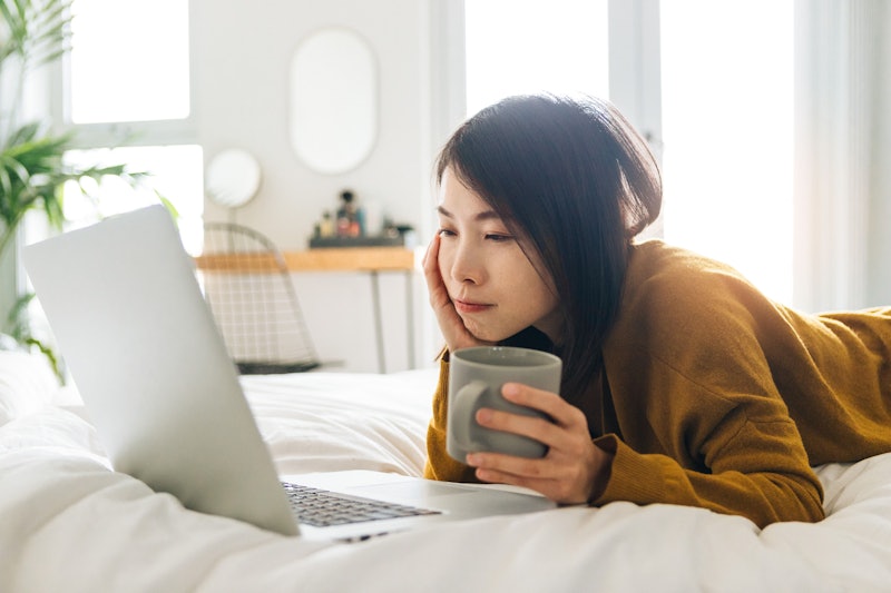 A woman looks at her laptop in bed with a cup of coffee. Here's how waking up later can affect your ...