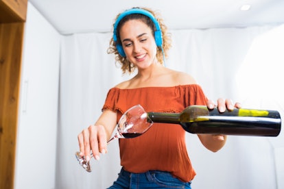 A young woman pours wine into a glass from a large bottle, while wearing bright blue headphones.