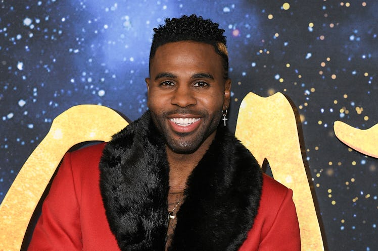 Jason Derulo smiles in front of a 'Cats' backdrop while wearing a red coat.