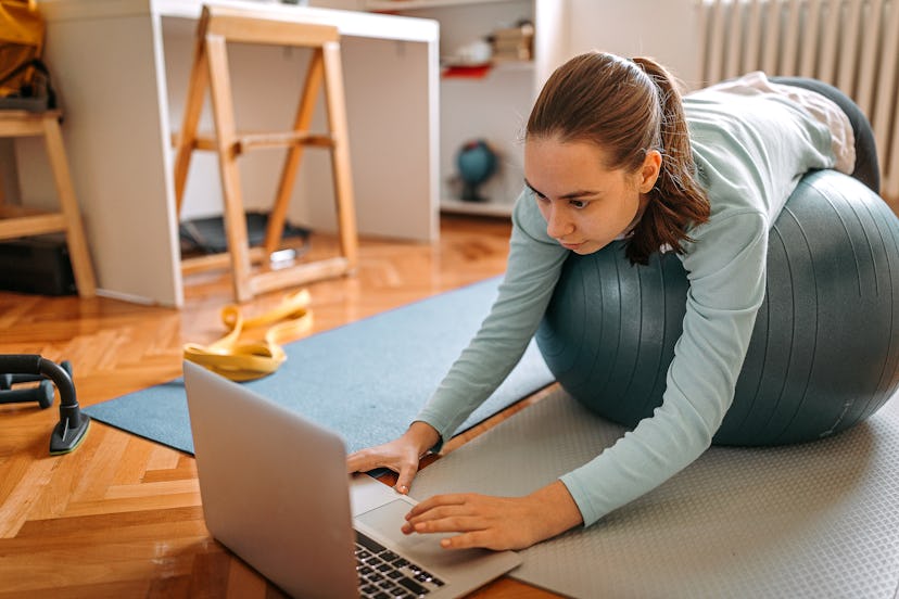 A person leans on her Swiss exercise ball while searching for a workout on her computer