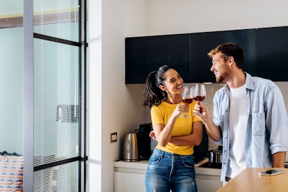 A young couple laughs in their kitchen while clinking their glasses of red wine.