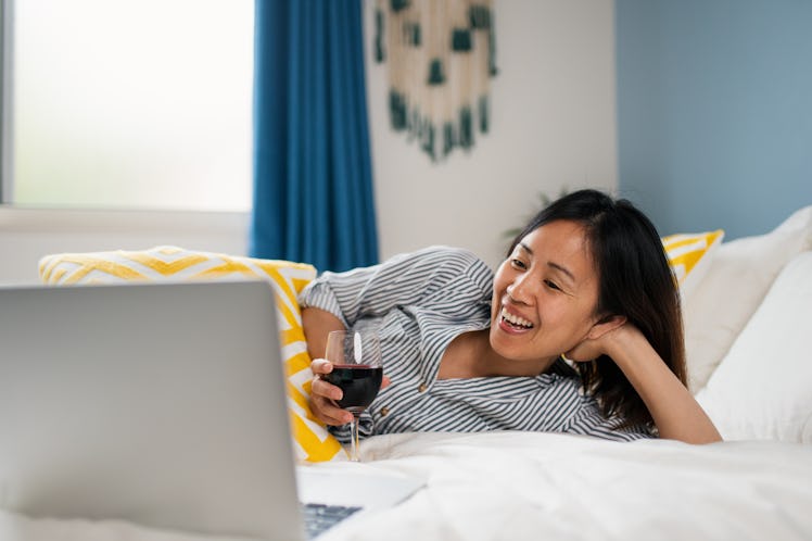 A young woman lays in bed and video chats her friends on her laptop while drinking red wine.