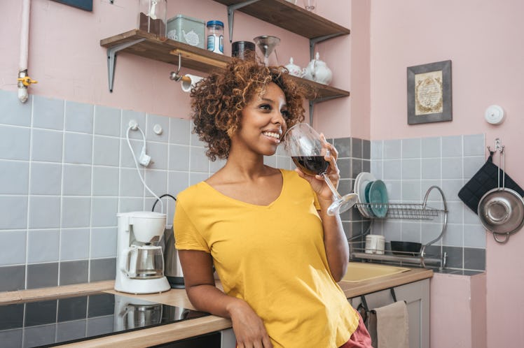 A young woman stands in her pink kitchen and drinks a glass of wine.