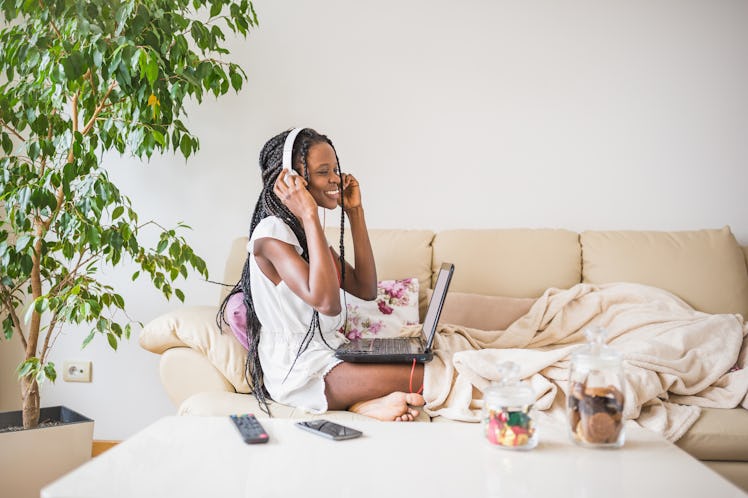 A young woman sits on her couch with her laptop and a pair of headphones, and is surrounded by blank...
