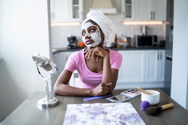 A young woman does a facial while sitting at her kitchen table with some makeup and a mirror.