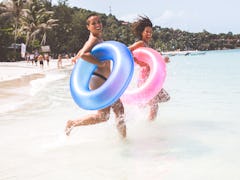 A young lesbian couples runs into the ocean with colorful tube floats in their hands.