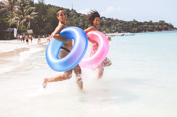 A young lesbian couples runs into the ocean with colorful tube floats in their hands.