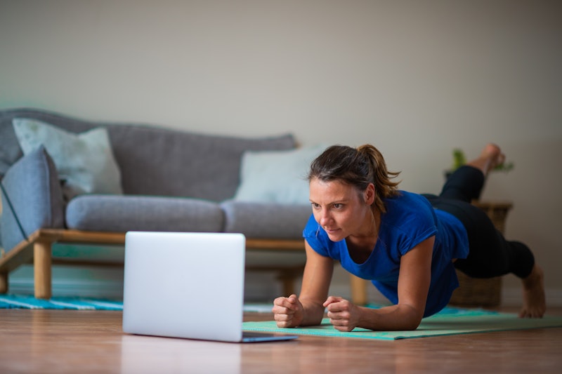 A person holds a plank in her living room while keeping an eye on the yoga class on her laptop. Taki...