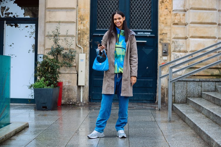 A young woman stands outside on a rainy street in the city in a tie-dye sweatshirt and jeans.