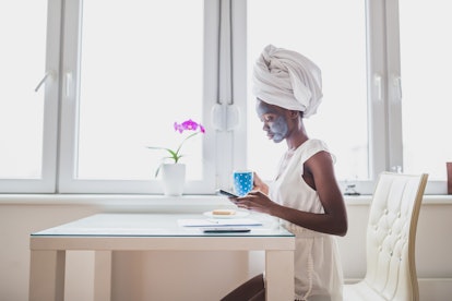 A woman with a face mask, her hair wrapped in a white towel, sitting while holding her phone and a b...