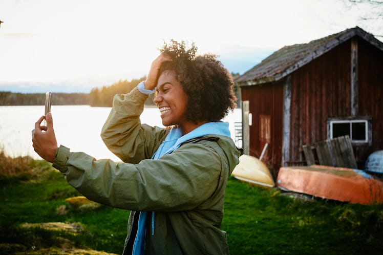 A young woman takes a selfie on her phone while standing next to a rustic home near a lake.