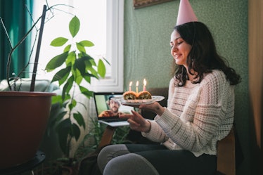 A young woman sits in a chair with a party hat on and a dessert with candles in her hand, and video ...