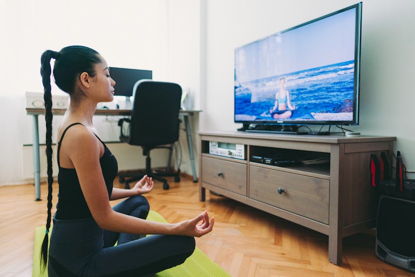 A person sits on their yoga mat and meditates in front of a yoga video on their television. Exercisi...
